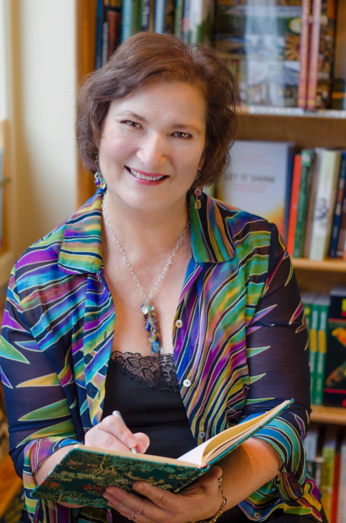 A woman wearing a colorful shirt sits in front of bookshelves, holding a pen and notebook, smiling warmly at the camera.