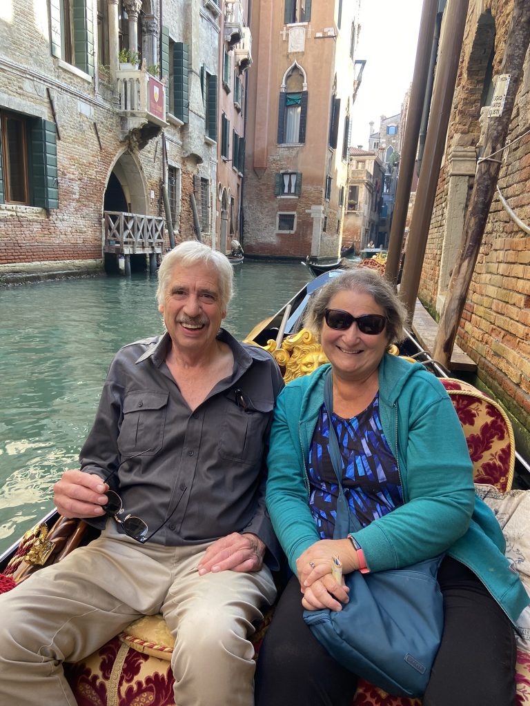 A couple smiling while seated in a luxurious gondola, gliding through the picturesque canals of Venice surrounded by historic architecture.