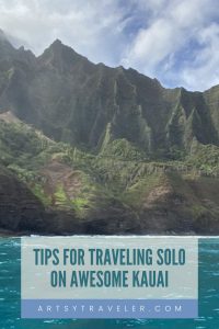 A scenic view of Kauai's lush, jagged mountains rising above the turquoise ocean under a partly cloudy sky. Overlaid text reads, "Tips for Traveling Solo on Awesome Kauai" with a footer that includes the website "artsytraveler.com."