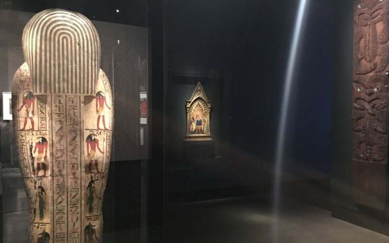 Exhibit at MONA: an ancient Egyptian sarcophagus adorned with intricate hieroglyphics and illustrations of deities, displayed in a dark exhibit space. A brightly lit ornate shrine is visible in the background.