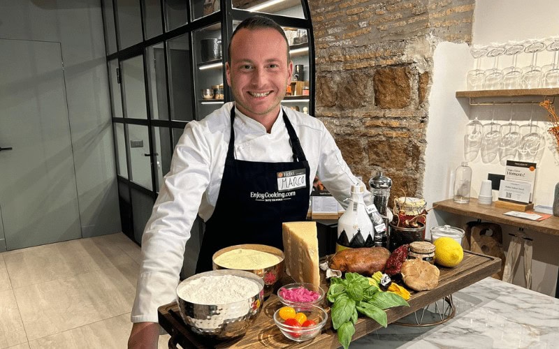 Chef Marco stands behind a table filled with fresh ingredients, ready to lead a cooking class in Rome."