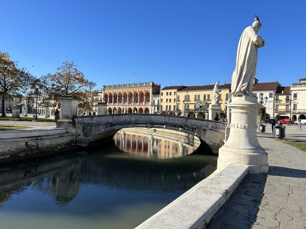 The iconic Prato della Valle in Padua, with its circular canal, white statues, and historic buildings in the background. A central attraction for visitors to Padua.