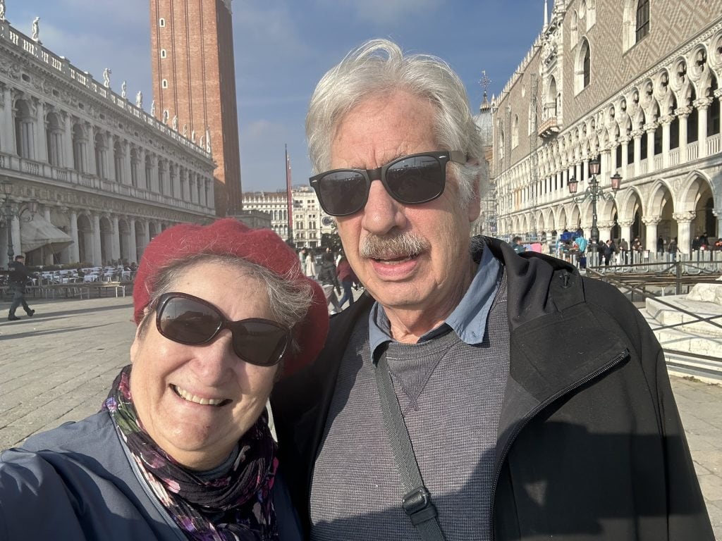 Author Carol Cram and artist Gregg Simpson standing in Saint Mark’s Square in Venice, surrounded by iconic Venetian landmarks. A perfect day trip from Padua to experience Venice's charm.