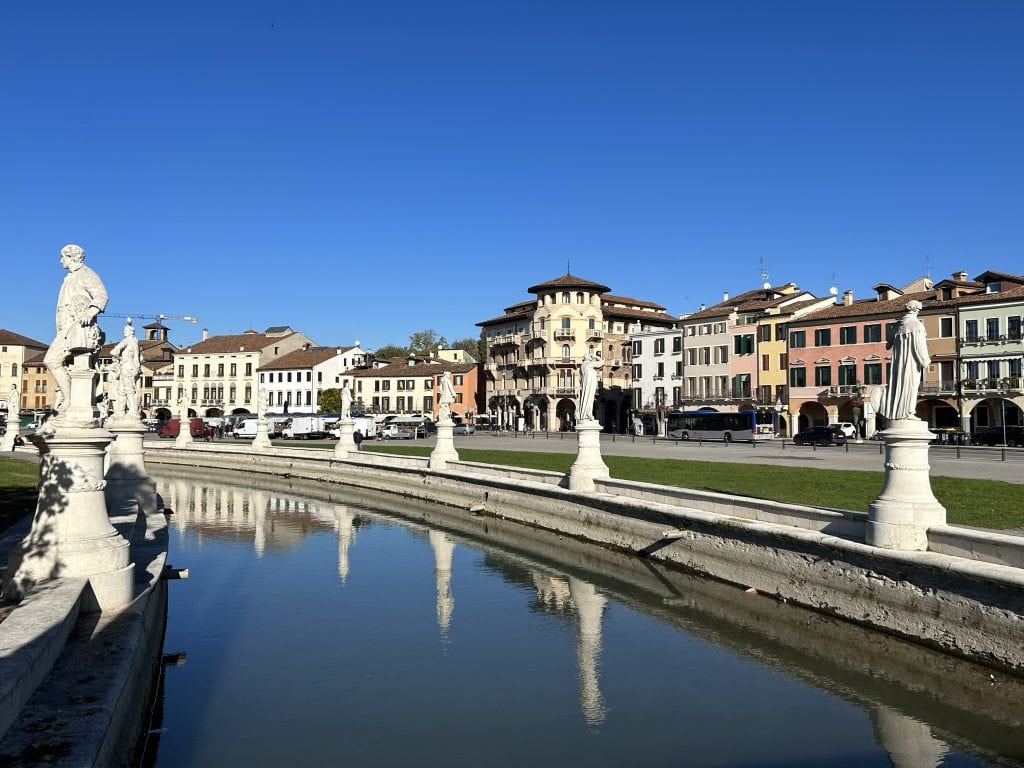 The iconic Prato della Valle in Padua, with its circular canal, white statues, and historic buildings in the background. A central attraction for visitors to Padua.