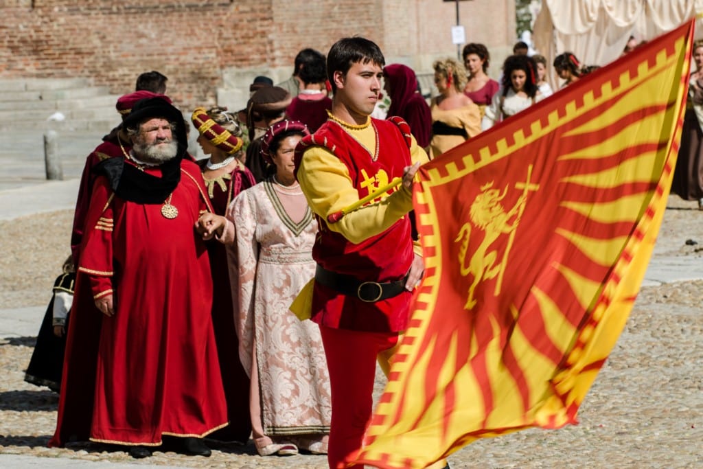 A man in a red and yellow medieval costume proudly waving a matching flag featuring a lion and cross emblem, with other people in period clothing walking behind him in a historical setting.