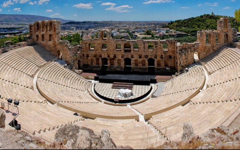 Panoramic view over the Odeon of Herodes Atticus on the Acropolis Slopes showing the sweep of seats and the backdrop of Roman-style ruins with the skyline of Athens beyond.