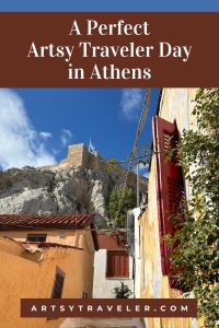 Pinterest graphic with the text A Perfect Artsy Traveler Day in Athens over a picture of a quaint village street in Athens with te view of the Acropolis beyound.