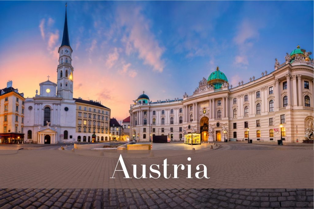 Panoramic view of Vienna’s Michaelerplatz at sunset, showcasing the neoclassical Hofburg Palace with green domes and the illuminated white St. Michael's Church steeple, under a sky filled with pink and purple hues. The cobblestone plaza is empty in the foreground.