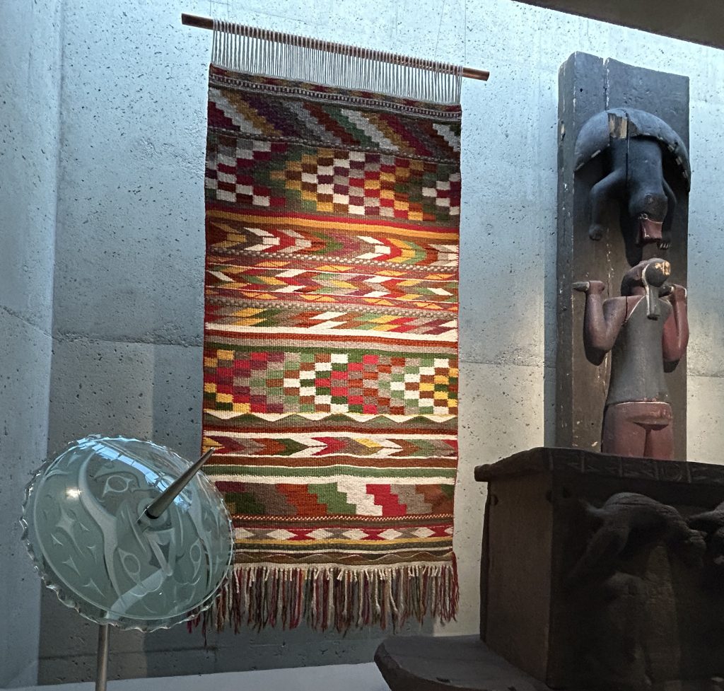 A vibrant woven blanket with intricate geometric patterns hanging in a museum exhibit, next to a wooden carving. These are featured on the way into the Great Hall at the Museum of Anthropology in Vancouver, Canada.