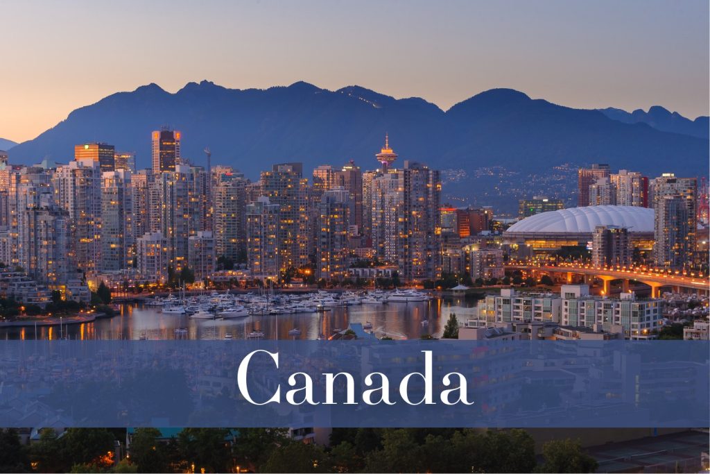 A panoramic view of Vancouver’s skyline at dusk, showing illuminated high-rise buildings, a stadium, and mountains in the background.