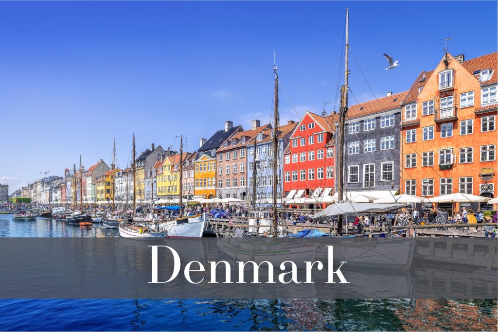 The colorful Nyhavn waterfront in Copenhagen, Denmark, featuring bright row houses, docked sailing boats, and bustling outdoor cafés, all reflected in the clear blue water under a cloudless sky.