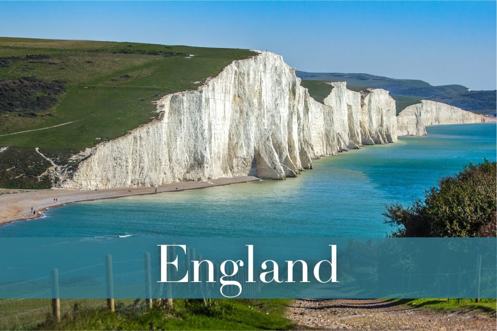 The iconic white chalk cliffs of the Seven Sisters in England, rising sharply along the coastline, with a turquoise sea lapping at the narrow beach and green hills rolling in the background.