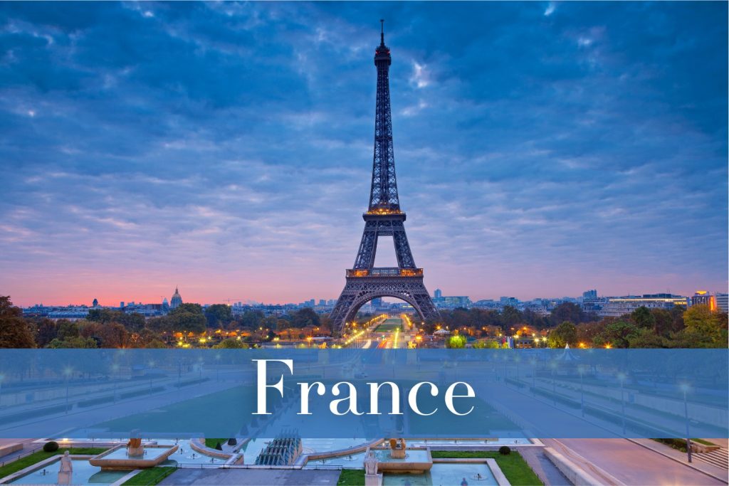 A twilight view of the Eiffel Tower in Paris, France, standing against a dramatic sky filled with soft clouds, with illuminated gardens and walkways in the foreground and the Paris cityscape stretching into the distance.