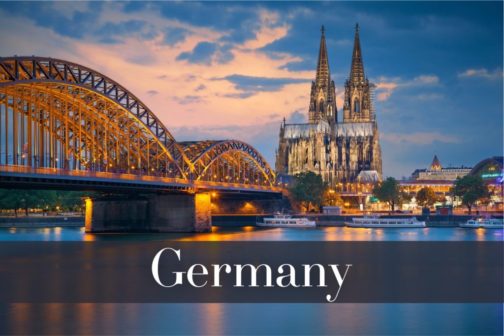 The Hohenzollern Bridge in Cologne, Germany, illuminated at night, with its steel arches glowing golden and reflecting on the Rhine River, alongside the towering Cologne Cathedral with its Gothic spires.