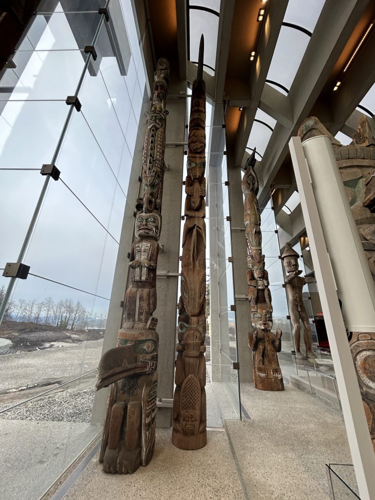 Tall Indigenous totem poles displayed in the Great Hall at the Museum of Anthropology museum with floor-to-ceiling glass windows and natural light.