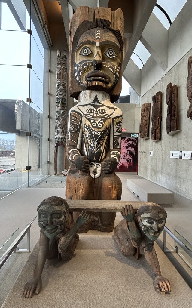 A large Indigenous wood carving featuring a figure with painted facial details, supported by two figures holding a wooden beam.