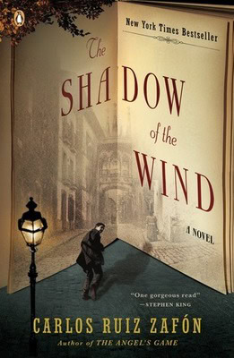 Book cover of The Shadow of the Wind by Carlos Ruiz Zafón. An open book forms the backdrop of a foggy, old street scene. A figure in a dark coat walks beneath a streetlamp. The title is displayed in red and black serif text.