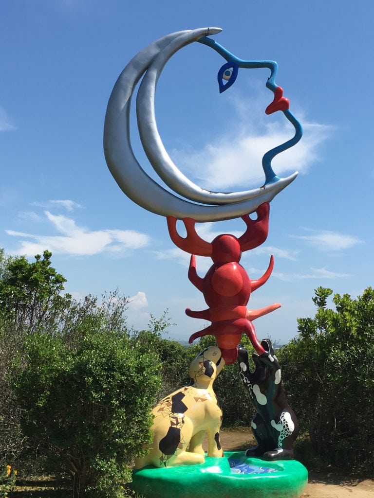 A whimsical sculpture from the Tarot Garden, representing a crescent moon forming a face, balanced atop a red figure resembling a lobster, supported by two animal-like figures on a multicolored base surrounded by greenery.