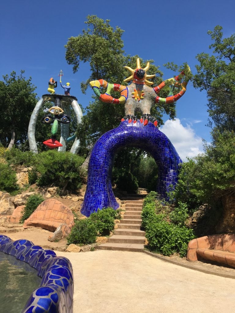 A vibrant mosaic sculpture of a fantastical figure from the Tarot Garden, featuring colorful, mirror-covered legs, outstretched arms, and a sun-like face, set amidst lush greenery under a bright blue sky.