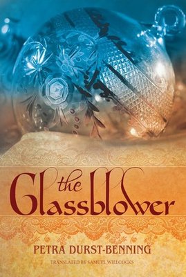 Cover of the book The Glassblower by Petra Durst-Benning, translated by Samuel Willcocks. The design features an ornate glass ornament with etched floral patterns, glowing softly in blue and gold hues. The title is written in elegant red script on a warm golden background.



