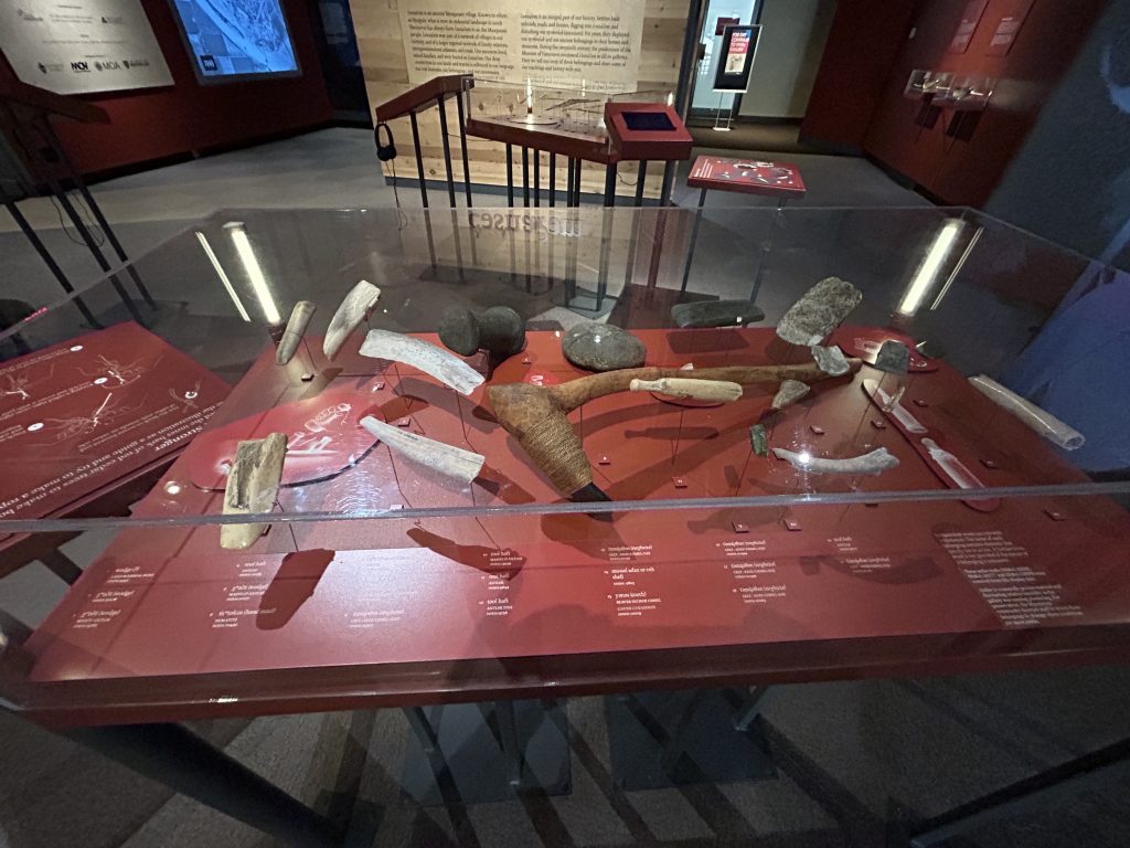 A close-up of historical artifacts, including stone tools, wooden items, and other indigenous implements, displayed in a glass case with detailed labels describing their origins and uses.