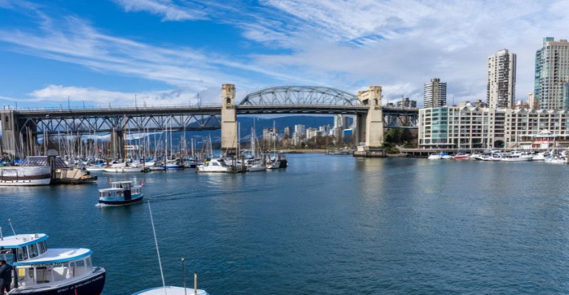 A wide-angle view of Burrard Bridge spanning a marina filled with boats. The city skyline and mountains are visible in the background, framed by a partly cloudy sky.