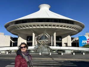 Carol Cram stands in front of the Museum of Vancouver, a futuristic looking white building with a saucer shaped roof and a fountain sculpture at the entrance. The building is labeled as the Museum of Vancouver, and the bright blue sky enhances the structure's architectural design.