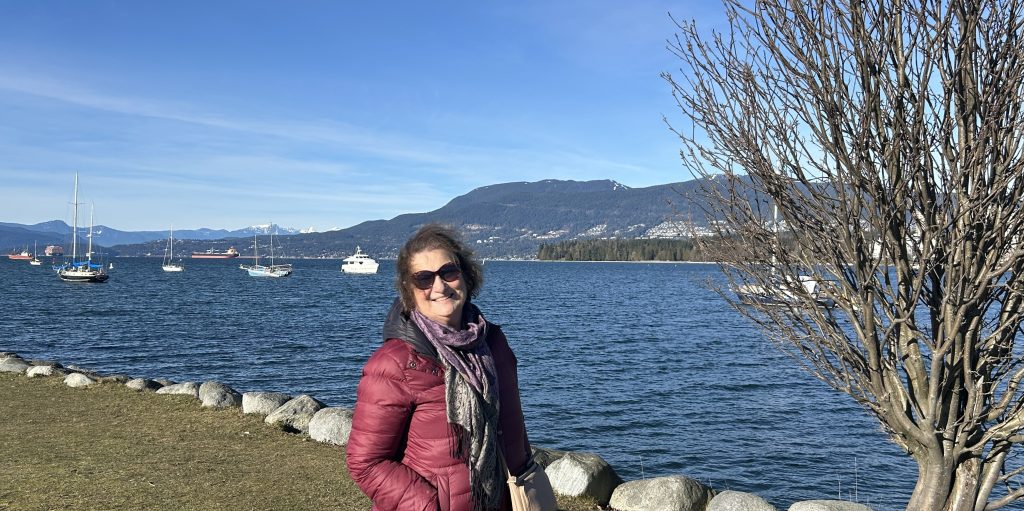 Carol Cram stands by a tree on the edge of a scenic waterfront. Sailboats float in the blue water, with mountains and clear skies in the background.