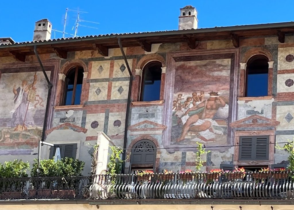 A historic building with elaborate Renaissance frescoes on the facade, including mythological and decorative elements, above a balcony with potted plants.