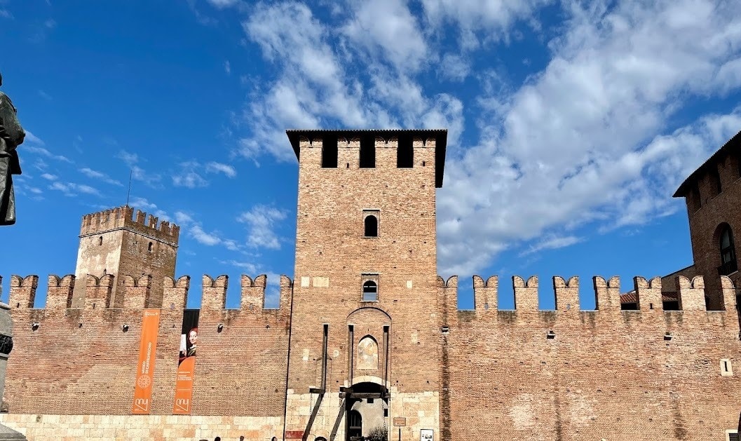 The brick facade of Castelvecchio, featuring crenelated towers and a central arched entrance, with visitors walking around the plaza in front.