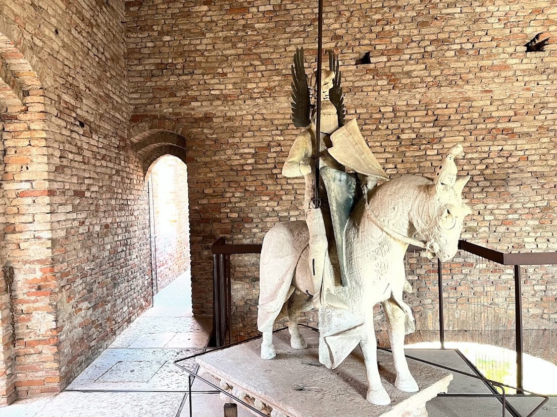 A medieval-style equestrian sculpture of a knight in armor, displayed against a brick wall with natural light streaming in.