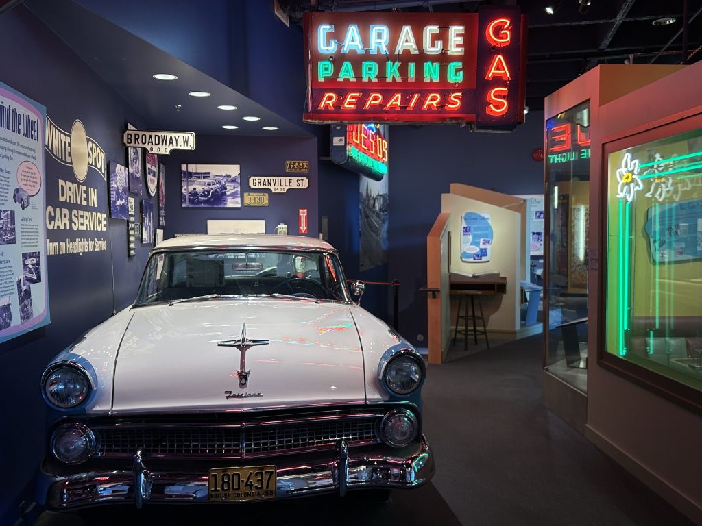 A retro museum exhibit featuring a classic 1950s white car parked under neon signs advertising garage, parking, and gas services. The setup evokes the mid-20th-century American automotive culture.