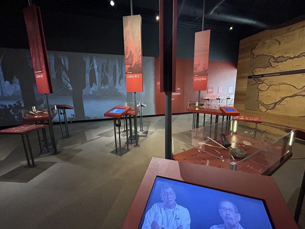 An exhibition showcasing First Nations artifacts and cultural information. The display includes red information panels with indigenous words and visuals, interactive screens, and historical tools arranged on tables under dim museum lighting.