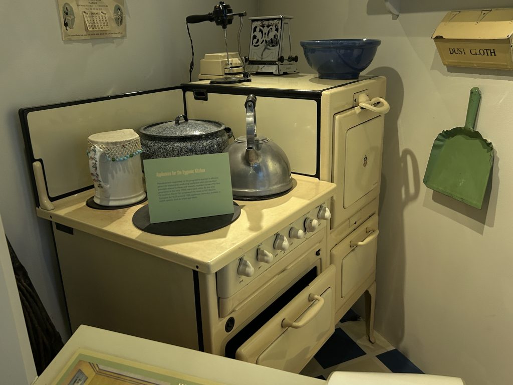 A vintage kitchen exhibit featuring an old-fashioned cream-colored stove, a kettle, and various household items like a dustpan and cloth, showcasing domestic life in the early 20th century.