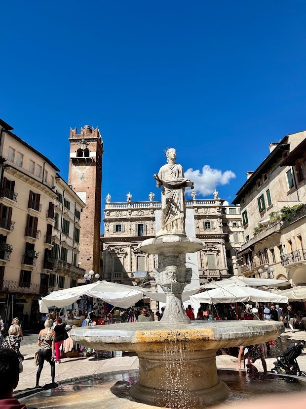 A central fountain in Piazza delle Erbe in Verona, featuring a statue of the Madonna di Verona atop a stone pedestal, surrounded by bustling market stalls and visitors.