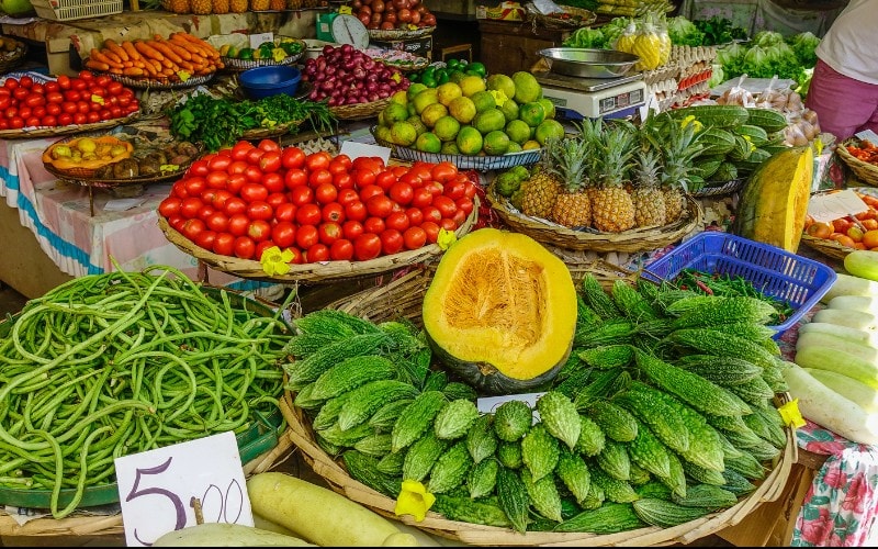 A vibrant market stall filled with colorful fresh produce, including tomatoes, pineapples, bitter melons, and long beans. Handwritten price signs are displayed, and the scene is bustling with bright colors.