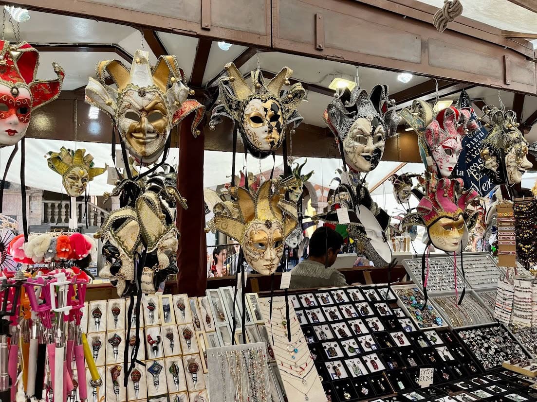 A market stall in Piazza delle Erbe, Verona, displaying an array of Venetian-style carnival masks with intricate designs in gold, red, and black. Below the masks are various trinkets, including keychains, jewelry, and other souvenirs, attracting visitors browsing the vibrant selection.