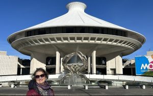 Carol Cram stands in front of the Museum of Vancouver, a futuristic looking white building with a saucer shaped roof and a fountain sculpture at the entrance. The building is labeled as the Museum of Vancouver, and the bright blue sky enhances the structure's architectural design.