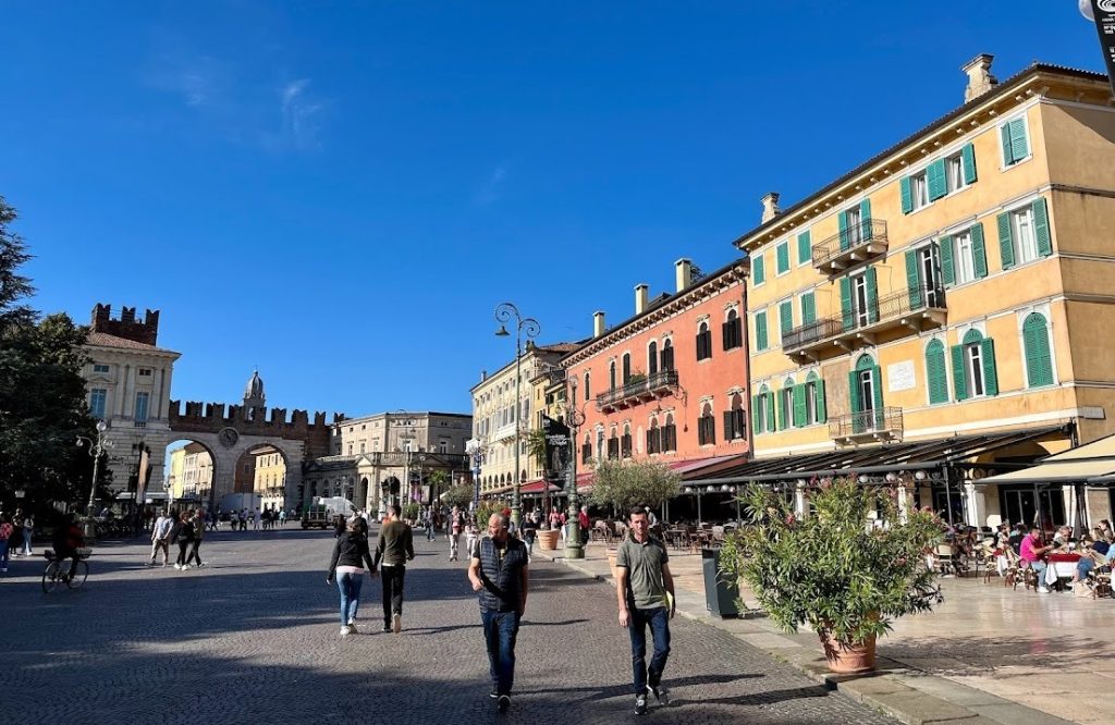 The piazza Bra in Verona with people strolling under the arches of the historic gate, flanked by colorful buildings and outdoor cafes.