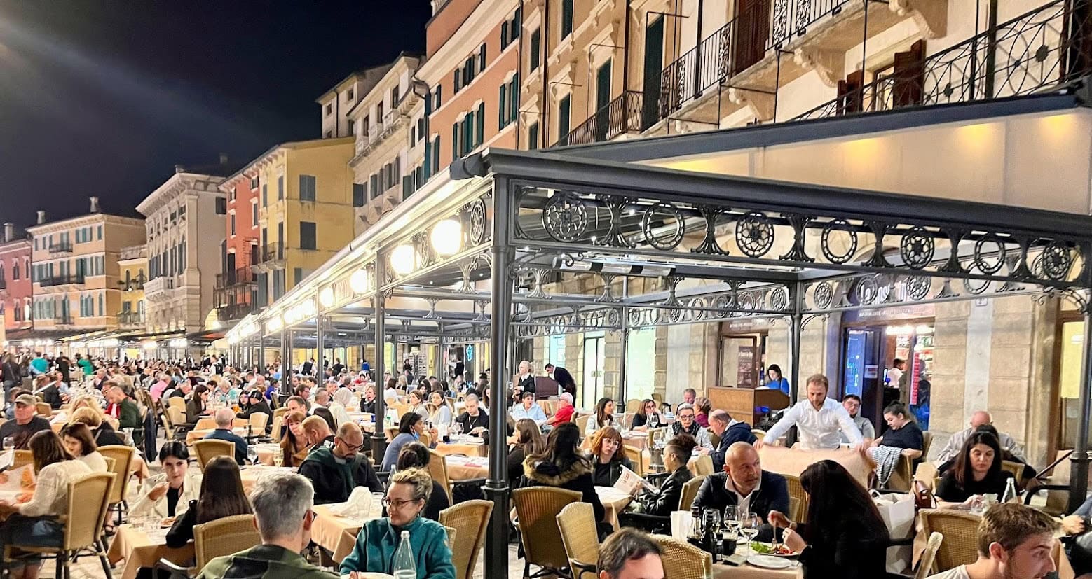 A bustling row of outdoor restaurants in Piazza Bra in Verona. The restaurants are packed with diners enjoying an evening meal beneath string lights and a covered terrace.