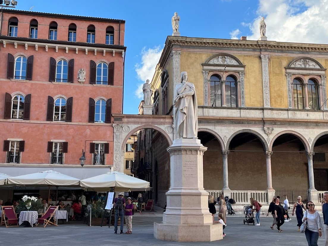 The Piazza di Signori in Verona with a statue of Dante at its center, surrounded by historic buildings, outdoor cafes, and visitors enjoying the open space.