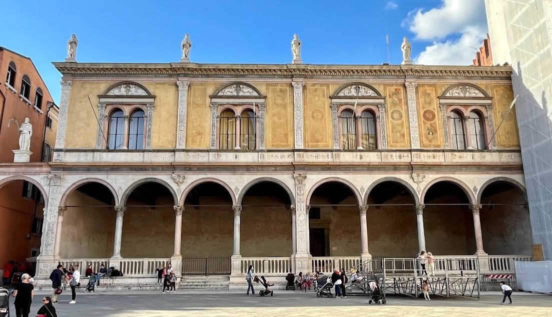 A historic loggia in Piazza dei Signori in Verona featuring arched colonnades and statues along its roofline, with visitors in the foreground.