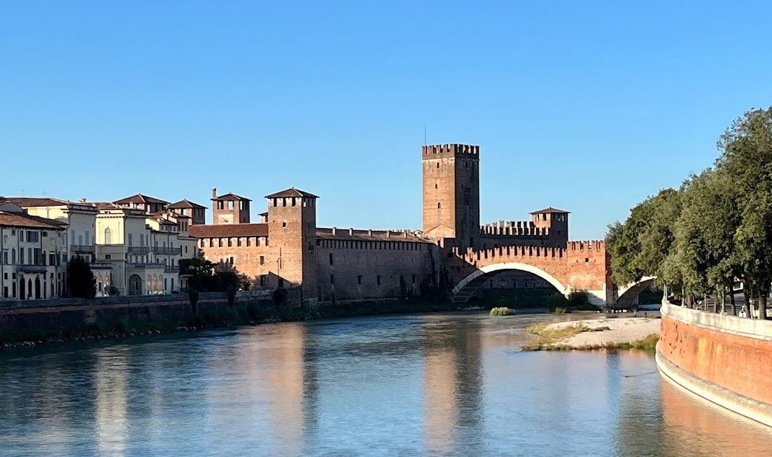 A scenic view of Castelvecchio Bridge and fortress reflecting on the calm waters of the Adige River under a bright blue sky.