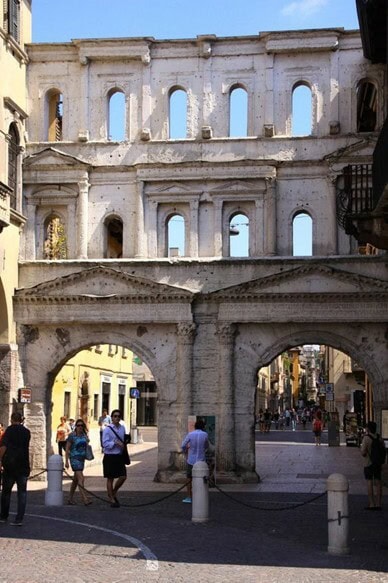 An ancient Roman gateway with arched entrances and upper stories featuring windows, situated at the entrance to a busy pedestrian street in Verona.