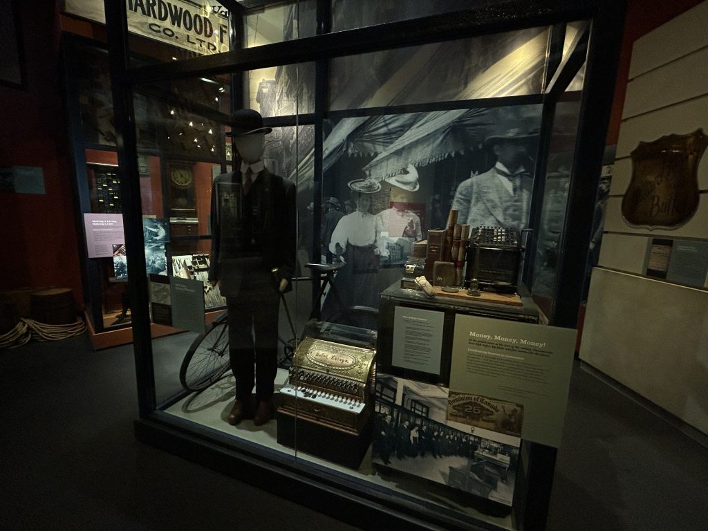 A museum display depicting pre-World War I commerce. Mannequins dressed in period clothing stand beside a vintage cash register and other business-related items in a glass-enclosed exhibit.