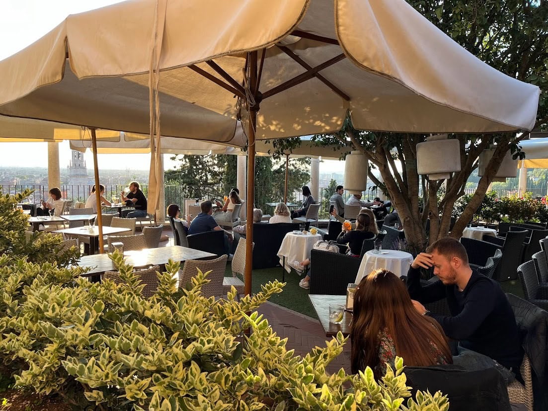 An outdoor terrace with shaded tables and chairs surrounded by greenery, offering diners a scenic view of the city at sunset.