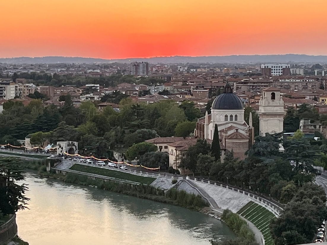 A panoramic view of Verona at sunset, showcasing the Adige River winding through the city and the dome of San Giorgio in Braida illuminated against the orange sky.