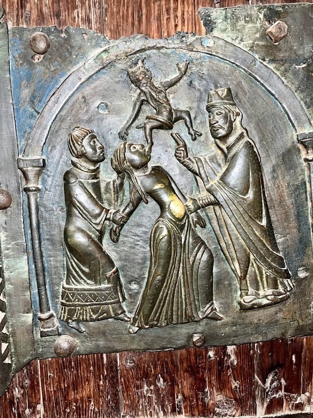 A detailed bronze relief panel depicting a dramatic scene of an exorcism, with a small demon being cast out from a woman by a figure in regal attire, while another figure supports her. The panel is framed by an arched design and shows intricate craftsmanship.