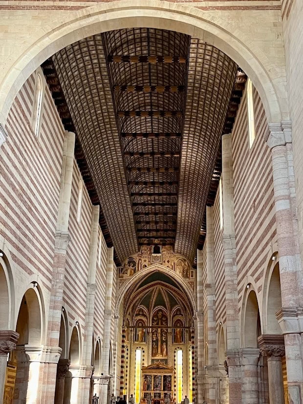 Grand interior view of the Basilica of San Zeno in Verona, showcasing its high vaulted ceiling, striped stone walls, and intricate altar at the far end.