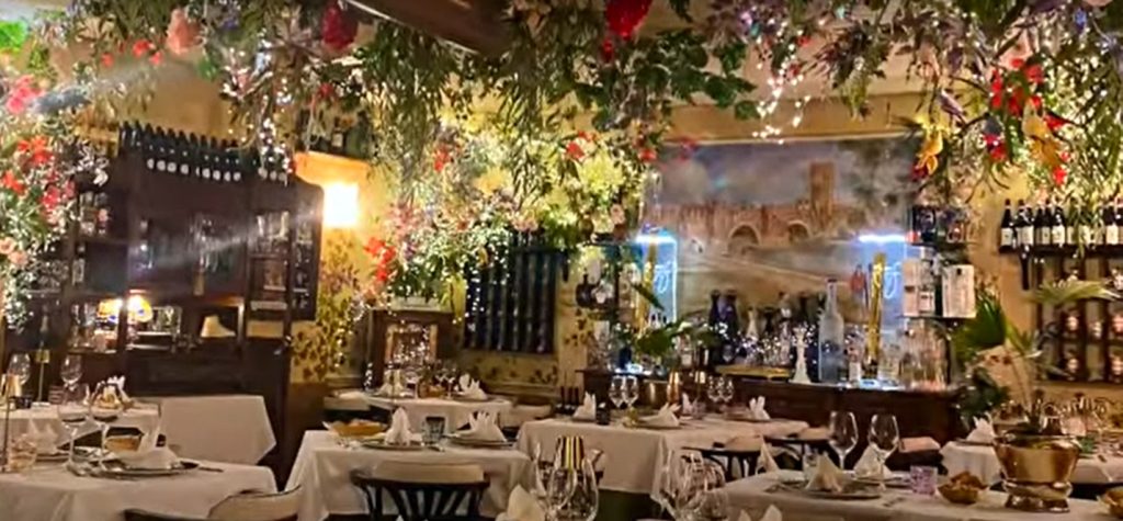 A cozy Italian restaurant with tables covered in white linens, surrounded by floral decorations and hanging greenery. Warm lighting and wine bottles enhance the romantic ambiance.